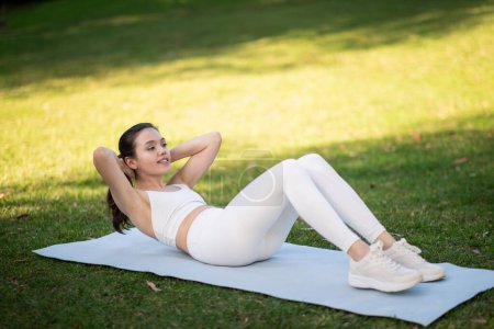 Photo for A smiling serene millennial caucasian woman engages in abdominal exercises on a yoga mat in a sunlit park, showcasing her fitness lifestyle in nature, outside, full length - Royalty Free Image