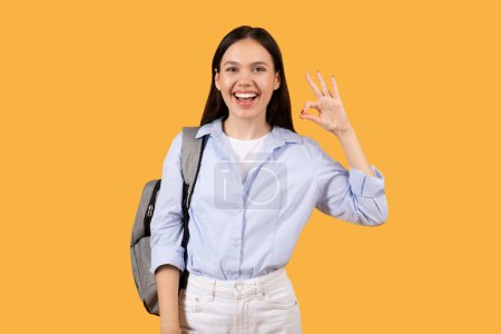 Confident lady in casual outfit with backpack giving okay sign, standing against vibrant yellow background, perfect for themes of approval, success, and student life