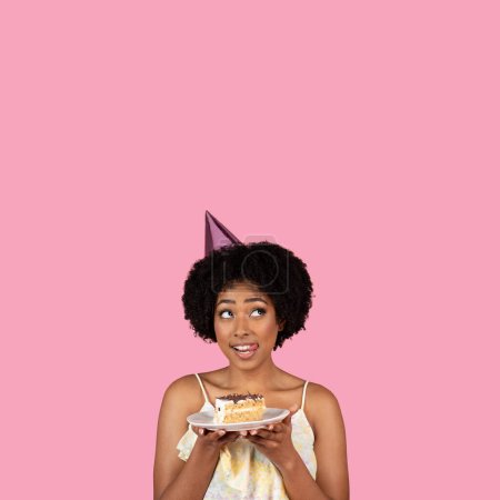 Téléchargez les photos : Une jeune femme heureuse étonnée dans un chapeau de fête, tenant une tranche de gâteau sur une assiette, lève les yeux grands ouverts sur un fond rose, exprimant surprise et plaisir, studio - en image libre de droit