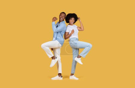 Téléchargez les photos : Joyeux couple afro-américain millénaire posant dos à dos avec des poings levés, en denim assorti et des chaussures blanches sur un fond jaune, studio, pleine longueur - en image libre de droit