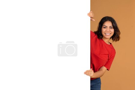 Foto de Mira esto. Smiling Arab Lady Stand By Copy Space On Blank White Advertisement Board, Positive Middle Eastern Woman Mostrando lugar libre para su diseño en la cartelera vacía - Imagen libre de derechos