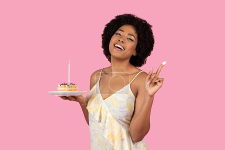 Téléchargez les photos : Une femme millénaire souriante rayonnante avec un sourire joyeux fait un vœu sur une bougie d'anniversaire allumée au sommet d'une délicieuse tranche de gâteau, son doigt ludique enduit de crème, profiter des vacances, studio - en image libre de droit