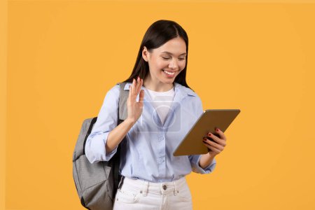 Photo for Engaged female student waves hello while focusing on her tablet, combining friendliness with technology against warm yellow backdrop, symbolizing e-learning - Royalty Free Image