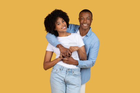 Téléchargez les photos : Heureux couple afro-américain millénaire en vêtements décontractés embrassant et souriant sur un fond jaune chaud, respirant l'amour et la compagnie, studio. Amour, relations - en image libre de droit