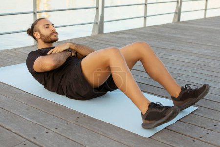 Seaside fitness. Motivated European guy in sportswear works on his abs doing crunches, exercises outdoors by the pier, showcasing healthy and active lifestyle. Full length