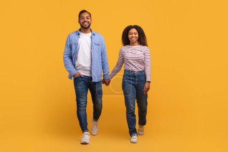 Téléchargez les photos : Portrait de heureux jeune couple noir tenant la main et marchant vers la caméra ensemble, homme et femme afro-américain posant sur fond de studio jaune, pleine longueur prise de vue avec espace de copie - en image libre de droit