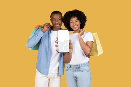 Téléchargez les photos : Couple afro-américain millénaire souriant affichant joyeusement un smartphone avec un écran vierge et tenant des sacs à provisions colorés, vente, sur un fond jaune vif, studio - en image libre de droit
