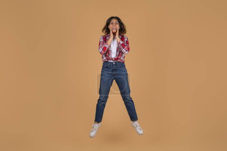 Photo for Emotional young middle eastern woman wearing casual outfit jumping in the air and touching her face, feeling excited, posing isolated on beige studio background, copy space - Royalty Free Image
