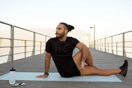 Summer fitness. Full length view of young man in activewear performing seated spinal twist asana outdoor, practicing yoga by the pier, achieving balance and wellbeing during session