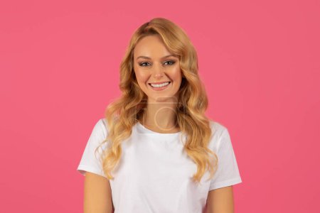 Téléchargez les photos : Belle dame blonde posant pour portrait sur fond de studio rose, rayonnant de confiance et de bonheur. Jeune femme souriant à la caméra. Concept de beauté et de mode - en image libre de droit