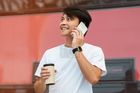 Photo for Closeup of millennial asian guy wearing white t-shirt drinking coffee and talking on phone with friend, standing on the street, looking aside at copy space - Royalty Free Image