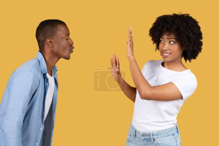 Téléchargez les photos : Homme souffle un baiser à une femme millénaire qui rejette ludique avec un geste de la main, tous deux habillés en vêtements décontractés sur un fond jaune, studio. Dites non et arrêtez le harcèlement - en image libre de droit