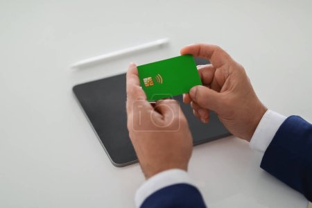 Foto de Primer plano de las manos de empresarios europeos de mediana edad examinando cuidadosamente una tarjeta de banco verde, probablemente verificando los detalles antes de hacer un pago, con una tableta y un bolígrafo en el fondo - Imagen libre de derechos