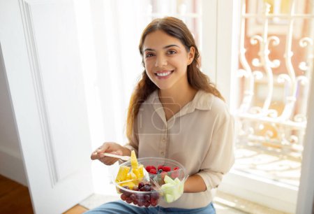 Photo for Happy young woman holding bowl of colorful mixed fruits and smiling at camera, beautiful european female enjoying healthy snack while sitting on floor by the window at home, closeup shot - Royalty Free Image