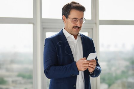 Photo for Engaged and focused european middle-aged man in a navy blue blazer using his smartphone with a gentle smile, standing by a window with natural light, chatting, use app in office - Royalty Free Image