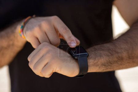 Photo for Unrecognizable athletic man in fitwear uses a smartwatch gadget for workout monitoring outdoors, checking his pulse during training, closeup of arms. Gym concept, motivation to fitness routine - Royalty Free Image