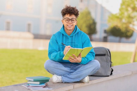 Photo for Happy brazilian student guy wearing eyeglasses learning taking notes in workbook, smiling to camera while studying seated with backpack at outdoor park setting near college building - Royalty Free Image