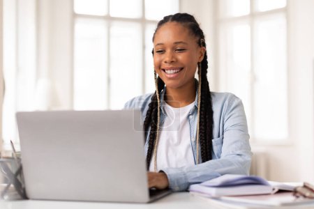 Téléchargez les photos : Étudiante noire joyeuse avec des tresses souriantes tout en utilisant son ordinateur portable pour l'apprentissage en ligne, assise au bureau blanc avec des cahiers et des lunettes de côté - en image libre de droit