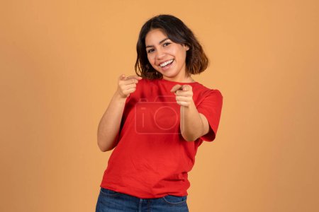 Photo for Joyful beautiful young arabic woman in red t-shirt pointing at camera and smiling, posing isolated on beige studio background. Middle eastern lady gesturing - Royalty Free Image