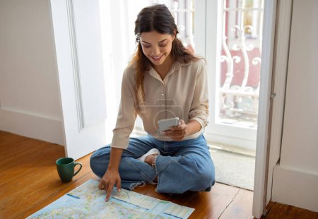 Photo for Young woman using her smartphone and city map to plan journey, sitting on wooden floor by the window at home, happy millennial female showcasing adventure preparation, getting ready for trip - Royalty Free Image