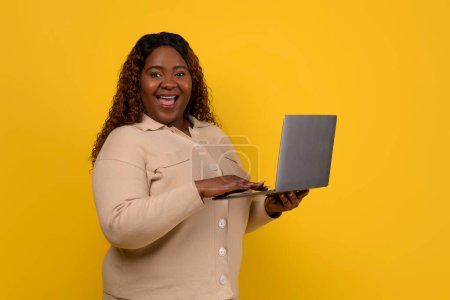 Photo for Cheerful overweight millennial black lady holding laptop computer looking at camera and exclaiming isolated on yellow studio background, checking online weigh loss course - Royalty Free Image