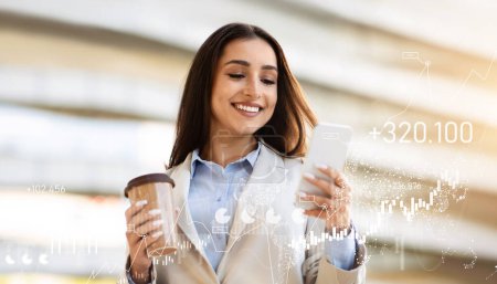 Téléchargez les photos : Femme d'affaires confiante avec une tasse de café sourit tout en utilisant son smartphone, debout dans un espace lumineux avec des graphiques numériques des marchés financiers superposant l'image. App pour le travail - en image libre de droit