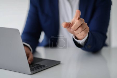 Téléchargez les photos : Un homme d'affaires européen en costume bleu pointe vers la caméra en travaillant sur son ordinateur portable à un bureau blanc, peut-être dans un geste de discussion ou d'explication. Travail, entreprise, démarrage - en image libre de droit
