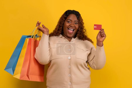 Contactless shopping. Excited chubby millennial black lady wearing beige shirt with colorful paper bags purchases and red plastic bank credit card posing on yellow studio background