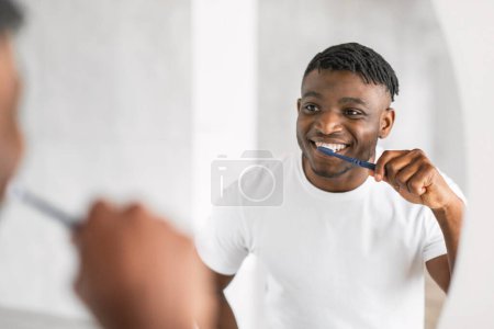 Foto de Guapo afroamericano millennial chico cepillarse los dientes con cepillo de dientes se encuentra junto al espejo, el cuidado de su salud bucal en el baño moderno. Rutina diaria de cuidado dental. Enfoque selectivo - Imagen libre de derechos
