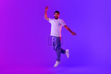 Téléchargez les photos : Joyeux jeune homme insouciant saute et lève le bras posant en plein air, dans des néons bleus et violets sur fond de studio, souriant à la caméra. Concept de bonheur, célébration des bonnes nouvelles - en image libre de droit