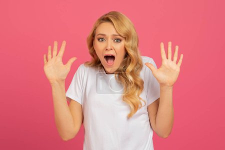 Téléchargez les photos : Femme blonde effrayée lève les mains dans un geste choqué sur fond rose, portant un t-shirt blanc décontracté, posant avec une bouche grande ouverte et une expression émotionnelle, photo de portrait en studio - en image libre de droit