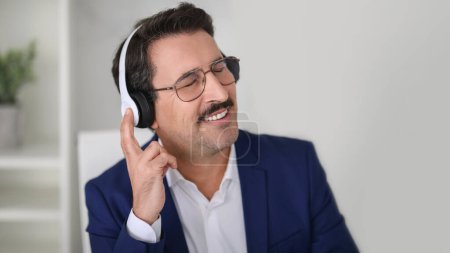 Photo for Smiling european middle aged man in a smart blue suit and glasses feeling the rhythm as he listens to music on his headphones, eyes closed in enjoyment, has fun, panorama - Royalty Free Image