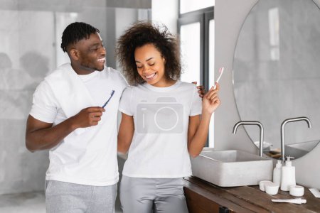Téléchargez les photos : Joyeux couple millénariste noir profite joyeusement des moments doux de la routine de soins dentaires, debout côte à côte avec des brosses à dents, brossant les dents le matin dans la salle de bain moderne à l'intérieur - en image libre de droit