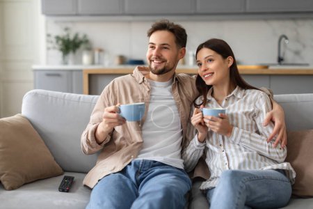 Entspannte und lächelnde Eheleute teilen gemütliche Momente mit Kaffeetassen auf bequemen Sofas im hellen Wohnzimmer, fühlen sich zufrieden und verbunden, genießen die Zeit am Wochenende