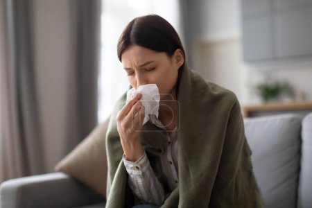 Mujer joven sintiéndose mal, envuelta en manta, se está sonando la nariz con tejido, lo que sugiere síntomas de resfriado o gripe mientras está sentada en el sofá en casa