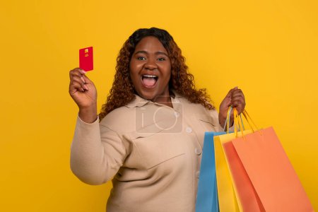 Photo for Happy pretty millennial overweight african american woman holding bunch of colorful paper bags purchases and credit card, enjoying shopping while season sale, isolated on yellow studio background - Royalty Free Image