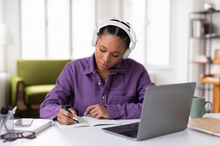 Engagierte schwarze Studentin mit Kopfhörern, die fleißig Notizen in Notizbuch von einer Online-Vorlesung auf ihrem Laptop macht, umgeben von Lernmaterialien