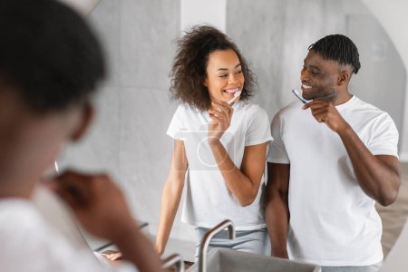 Foto de Pareja afroamericana se para en el baño cepillándose los dientes para el cuidado dental y el bienestar, sonriéndose mutuamente, disfrutando de su amor y su rutina de autocuidado saludable. Enfoque selectivo - Imagen libre de derechos