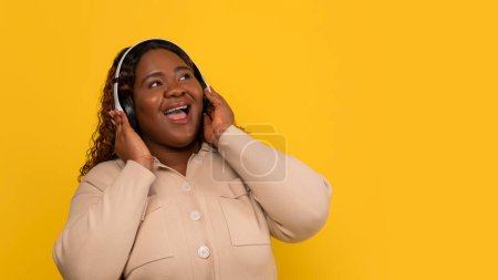 Photo for Positive pretty long-haired overweight millennial african american woman listening to her favorite song and looking at copy space, using wireless headphones, yellow background, panorama - Royalty Free Image