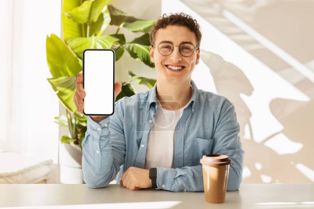 Photo for Cheerful caucasian young man student with curly hair and glasses showing a smartphone screen, ready for a presentation, with a takeaway coffee cup on a sunlit table. Recommendation app - Royalty Free Image
