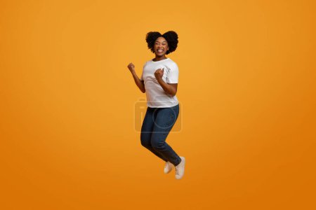 Foto de Emocionado joven afroamericana dama saltando en el aire sobre fondo de estudio naranja, apretando los puños y sonriendo, celebrando el éxito, longitud completa, espacio de copia - Imagen libre de derechos