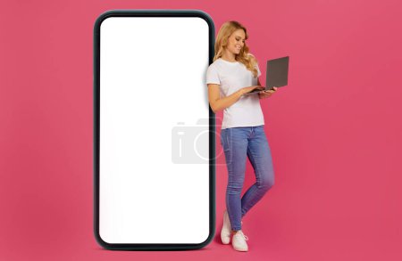 Téléchargez les photos : Freelance numérique. Blonde jeune femme websurfing sur ordinateur portable, tout en se tenant près d'un grand téléphone portable avec écran blanc, sur fond rose en studio. Maquette pour site web ou application - en image libre de droit