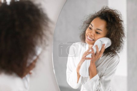 Photo for Happy black millennial lady wiping her face with soft towel in modern bathroom, standing in silk bathrobe and smiling to her reflection in mirror. Female skincare concept - Royalty Free Image
