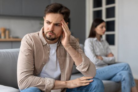 Photo for Upset man with troubled expression holding his head in his hand, foreground focus, with disheartened woman sitting arms crossed behind him - Royalty Free Image