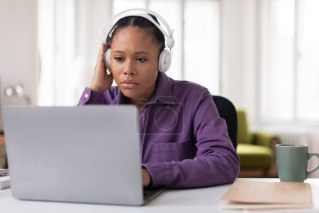 Photo for Thoughtful female student wearing headphones appears focused as she uses her laptop, possibly for studying or attending an online class at home - Royalty Free Image