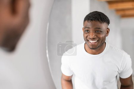Téléchargez les photos : Jeune homme noir en t-shirt blanc se prépare pour la journée dans sa salle de bain à la maison, souriant à son reflet dans le miroir. routine de toilettage quotidien masculin de l'hygiène et des soins de la peau. Concentration sélective - en image libre de droit