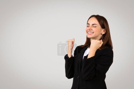 Téléchargez les photos : Jeune femme d'affaires caucasienne extatique en costume noir chic pompe ses poings dans les airs avec un sourire joyeux, célébrant une victoire personnelle ou professionnelle, isolé sur fond gris, studio - en image libre de droit