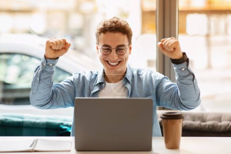 Joyful caucasian young man student with curly hair wearing glasses and a blue shirt celebrates success in front of his laptop at a bright cafe with a takeaway coffee cup, enjoy win