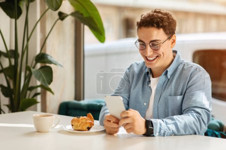 Téléchargez les photos : Heureux jeune homme caucasien étudiant avec les cheveux bouclés et des lunettes en utilisant son smartphone à une table de café avec un croissant frais et café, profiter d'une matinée lumineuse à l'intérieur. Concevoir pour le travail - en image libre de droit