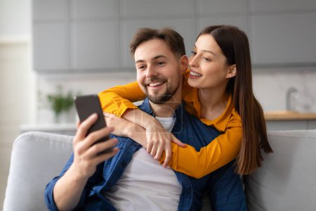 Téléchargez les photos : Souriant jeunes conjoints en tenue décontractée profiter du moment ensemble, capturer selfie sur téléphone portable tout en embrassant sur un canapé confortable à la maison - en image libre de droit
